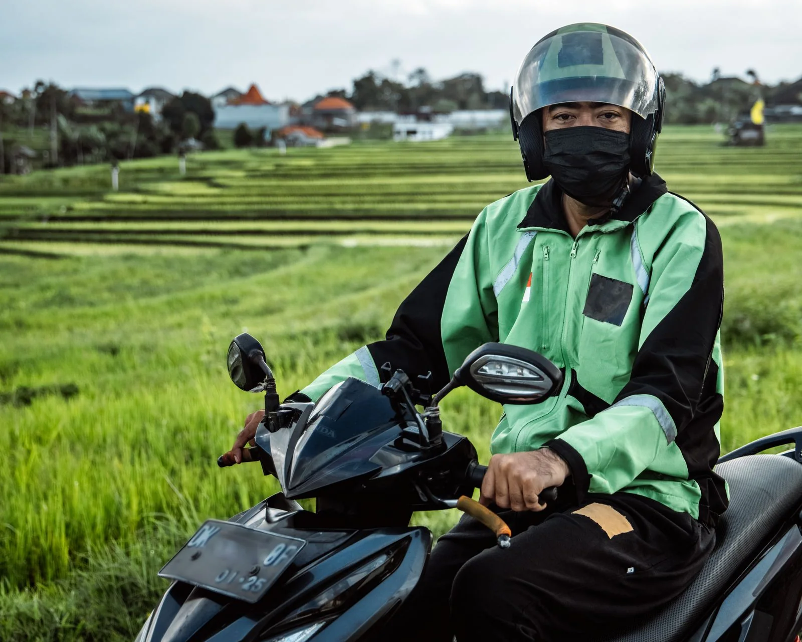 A delivery rider in a green jacket and helmet on a scooter beside green fields