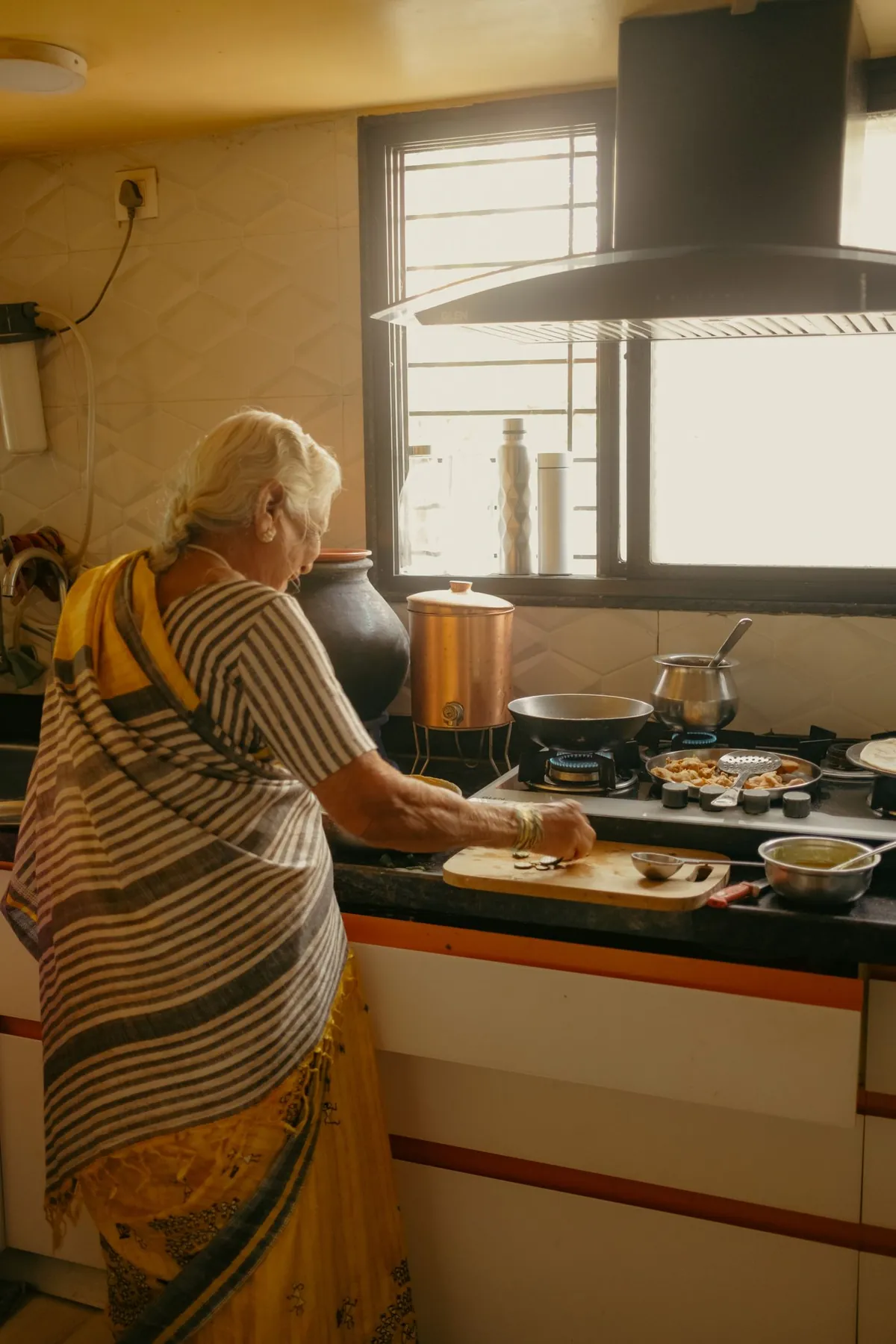 A woman in a sari cooking at the stove in a sunlit Indian home kitchen